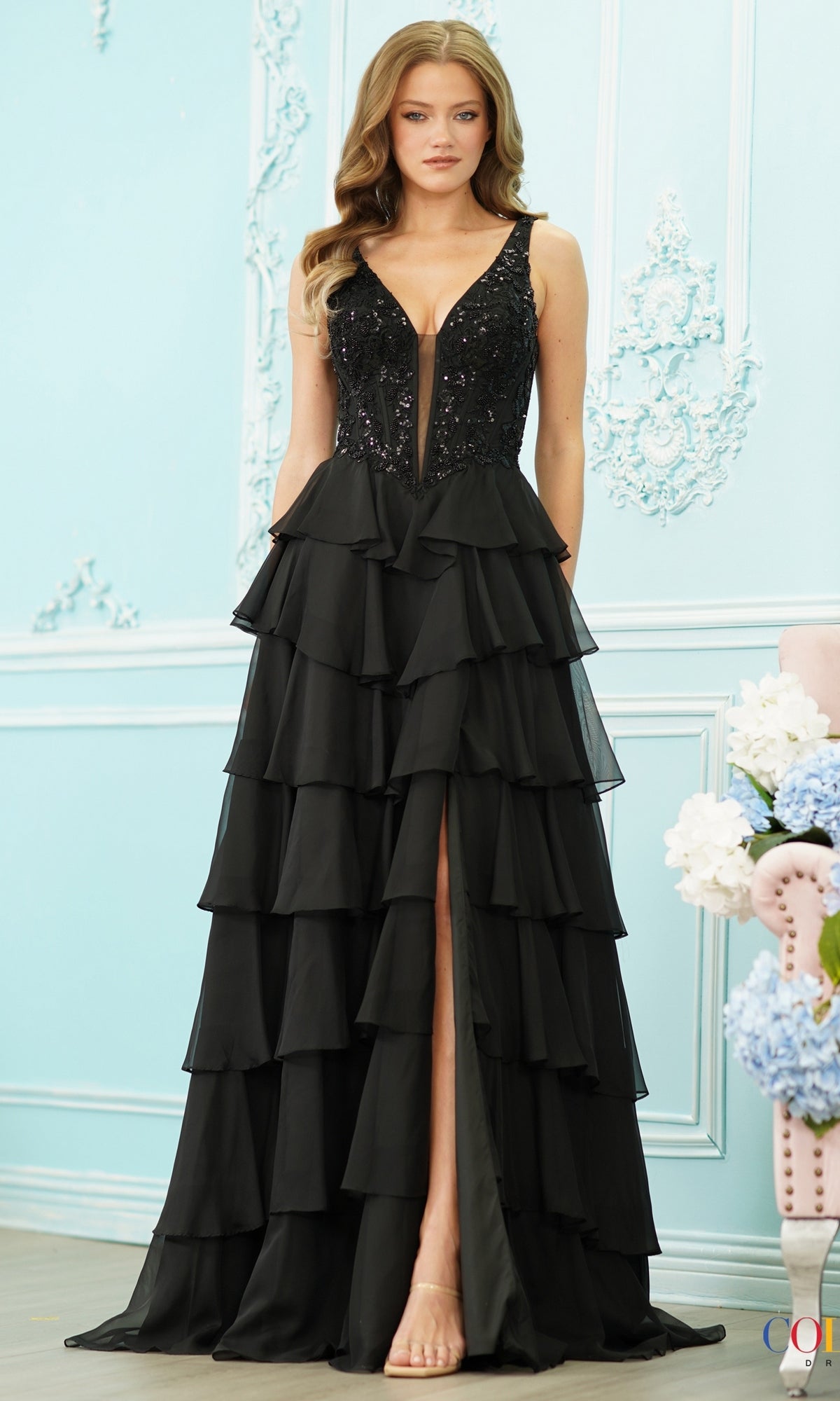 A woman wearing a black Long Prom Dress: Colors Dress 4177 featuring a V-neck, sequined bodice, tiered ruffled skirt, and a high front slit.